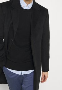 Mann in einem schwarzen Mantel, schwarzem Pullover, hellblauem Hemd mit Kragen und blauen Hosen, mit einer Hand in der Tasche.