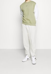 Person som bär en ljusgrön och vit långärmad sweatshirt, off-white sweatpants och vita sneakers med svarta Nike-logotyper, stående mot vit bakgrund.