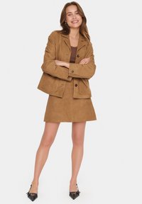 Blazer en daim marron avec une mini jupe assortie, présentant un col cranté, une fermeture à boutons et des lignes épurées. Des escarpins noirs à bout pointu complètent la tenue.
