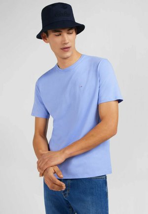 T-shirt en coton bleu clair à manches courtes, col rond, avec un petit logo sur la poitrine, associé à un jean bleu et un chapeau seau noir.