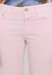 Gros plan sur un pantalon en denim rose clair avec un bouton argenté et des poches avant, porté avec une chemise colorée rentrée.
