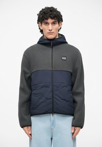 Fleece bunda v tmavošedej a námorníckej modrej, s uzatváraním na zips, kapucňou a prešívanou textúrou na dolnej časti. Logo značky na hrudi.