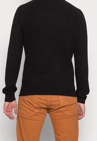 Schwarzer Rippensweater mit langen Ärmeln, Rundhalsausschnitt und strukturiertem Finish, kombiniert mit orange-braunen Hosen mit Gesäßtaschen.