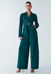 Teal long-sleeve jumpsuit with a belted waist, structured collar, wide legs, and smooth texture. Features gold hardware on the belt.