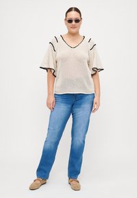 Top de malha bege com mangas largas, decorado em preto, combinado com jeans de perna reta azul claro e sapatos bege. Textura suave, trama aberta.