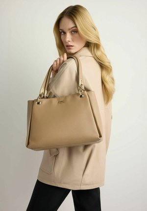 Blondynka w beżowej kurtce trzyma dużą jasnobrązową skórzaną torebkę ze złotymi elementami na ramieniu na jasnym tle.