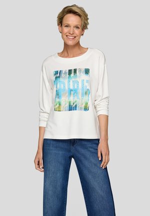 Weißes Langarmshirt mit einem bunten Grafikdruck in Blau-, Grün- und Weißtönen. Kombiniert mit locker sitzenden blauen Jeans.