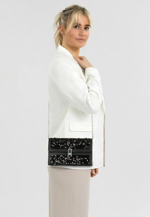 Sac à main noir en sequins avec une bandoulière chaîne, forme rectangulaire, finition brillante et un fermoir argenté. Porté avec une tenue neutre.
