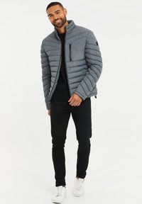 Grå pufferjakke med horisontale sømme, høj krave, lynlås lomme på venstre bryst, båret med sorte bukser og hvide sneakers.