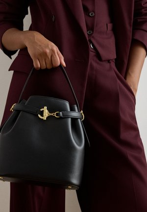 Bordó öltönyt viselő személy fekete bőr kézitáskát tart arany színű kapoccsal és felső fogantyúval az oldalán.