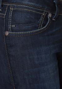 Vaqueros de mezclilla azul oscuro con una textura suave, que presentan remaches, un pequeño bolsillo frontal y costuras amarillas a lo largo de las costuras.