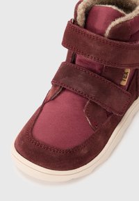 Bota de tornozelo em bordeaux com materiais em camurça e tecido, apresentando duas tiras de velcro, forro em pelúcia e sola de borracha texturizada.
