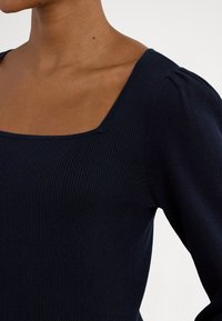 Tmavomodrý rebrovaný top s dlhými rukávmi, štvorcovým výstrihom a nafúknutými rukávmi, ktorý ukazuje jemnú textúru a priliehavý dizajn.