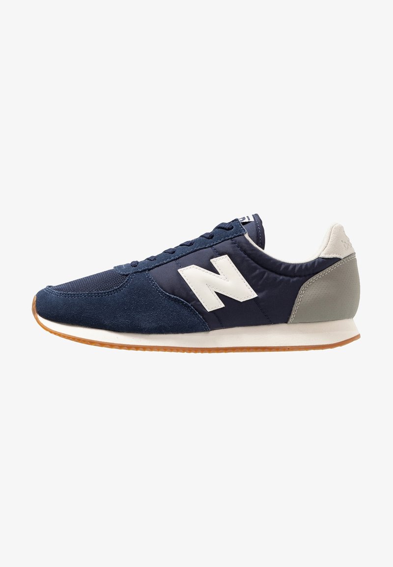 Baskets de sport bleu marine et gris avec logo blanc "N", fermeture à lacets et semelle en caoutchouc naturel, vue de côté sur fond blanc.