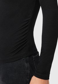 Top lungo a maniche ribattute nere con arricciature laterali e design aderente, abbinato a jeans in denim scuro. Texture liscia e cuciture pulite.