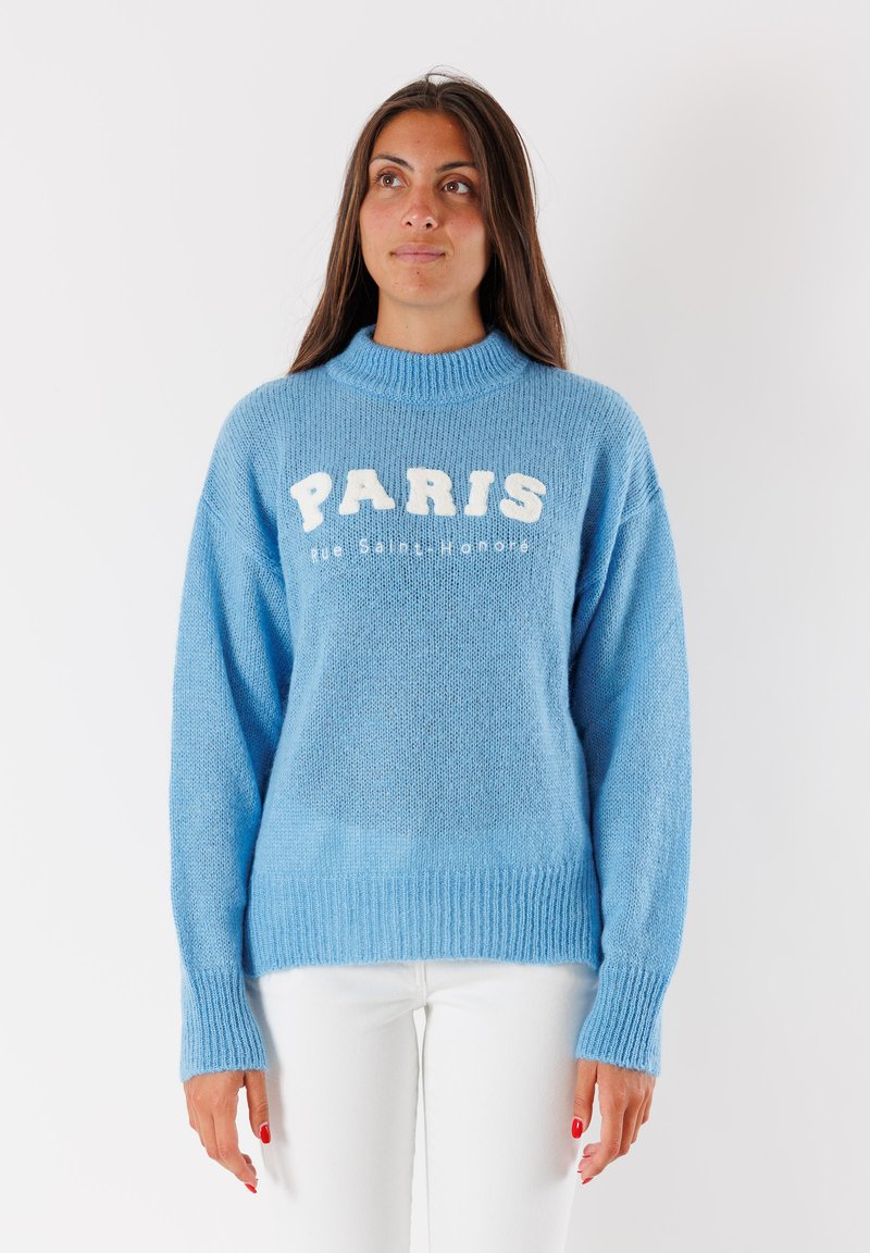 Donna con lunghi capelli castani che indossa un maglione lavorato a maglia di colore azzurro chiaro con la scritta "PARIS Rue Saint-Honoré" e pantaloni bianchi, in piedi su uno sfondo neutro.