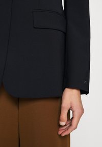 Blazer bleu marine à la texture lisse, avec une poche passepoilée simple et un détail de poignets boutonnés, associé à un pantalon marron en tissu contrastant.