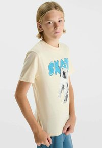 Jeune personne blonde portant un t-shirt beige avec le texte "SKATE" et un motif d'ours polaire tenant un skateboard à damier.
