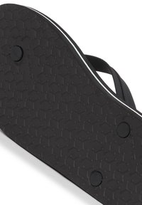 Svart flip-flop sula med ett texturerat hexagonalt mönster. Har runda, svarta accentknappar och en tunn vit rand längs kanten.