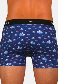 Boxers bleu marine avec un motif répété de poissons bleus et de flèches rouges. Ceinture noire avec le logo "WAXX". Matière douce et extensible.
