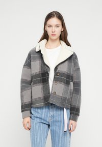 Jachetă croptă în carouri gri, cu un guler alb pufos. Țesătură moale și texturată și trei nasturi maro. Asortată cu blugi dungi în nuanțe deschise de albastru.