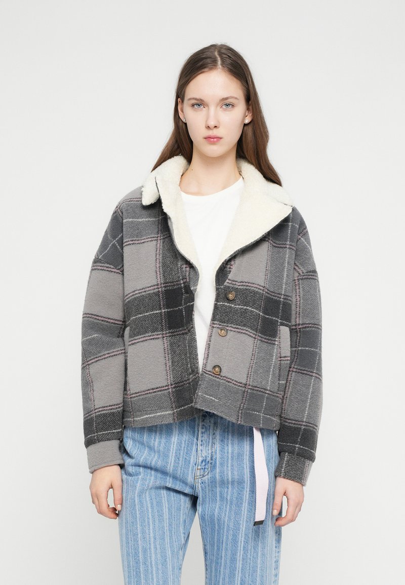 Jachetă croptă în carouri gri, cu un guler alb pufos. Țesătură moale și texturată și trei nasturi maro. Asortată cu blugi dungi în nuanțe deschise de albastru.