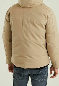 Beżowa kurtka puchowa z kapturem, z pikowanym wzorem i teksturowaną powierzchnią; noszona na ciemnych dżinsach z detalami na tylnych kieszeniach.