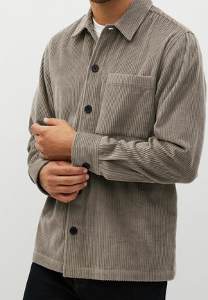 Mand justerer manchet på beige fløjlsnederdel med brystlomme, lagdelt over hvid undertrøje, mod ensartet baggrund.