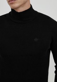 Schwarzer Pullover mit hohem Kragen, aus Rippstoff gefertigt. Verfügt über ein dezentes Logodetail auf der Brust. Die taillierte Passform betont die Silhouette.