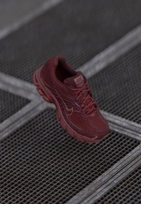 Červená bežecká obuv Nike s sieťovaným zvrškom, textúrovanými detailmi, polstrovaným golierom a hrubou podrážkou, umiestnená nad mriežkovaným povrchom.