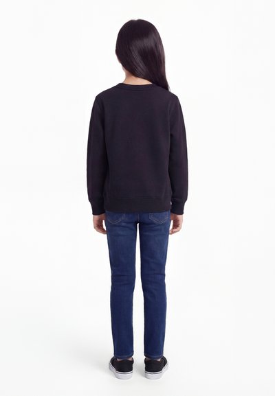 Dieťa s dlhými tmavými vlasmi, oblečené v čiernej mikine, modrých rifle a čiernych teniskách, stojace otočené chrbtom na bielom pozadí.