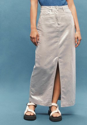 Kvinde iført en lys beige lang nederdel med en frontslids og hvide sandaler, stående imod en ensfarvet blå baggrund.