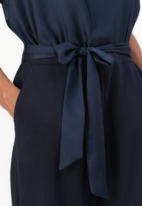 Tessuto blu navy con una texture liscia, caratterizzato da una cintura annodata in vita e tasche laterali. Il design è semplice ed elegante.