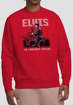 Person trägt einen roten Pullover mit einem Bild von Elvis Presley, der auf der Bühne Gitarre spielt, vor einer Menschenmenge und dem weißen Text "'68 Comeback Special".