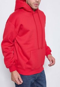 Homme portant un sweat à capuche rouge vif avec cordons et poche avant, associé à un jean bleu, sur un fond clair.