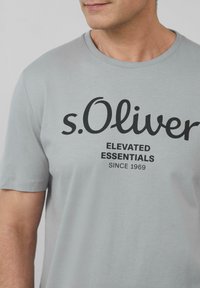 Jasnoszara bawełniana koszulka z krótkim rękawem i okrągłym dekoltem, z czarnym napisem "s.Oliver Elevated Essentials Since 1969".