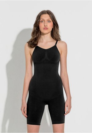 Mlada žena s valovitom smeđom kosom, odjevena u crni uski bodysuit s tankim naramenicama, stoji nasuprot jednostavnoj svijetloj pozadini.