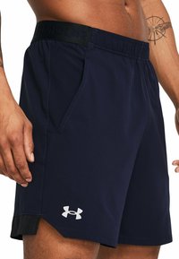 Mann mit bloßem Oberkörper, der marineblaue Under Armour Sportshorts mit schwarzem Bund und Logo am linken Bein trägt, Tattoo am rechten Arm sichtbar.