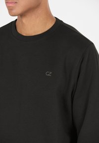 Svart bomullströja med ribbad rund hals och subtil "CZ" logotyp på bröstet. Slät textur, minimalistisk design, figursydd silhuett.