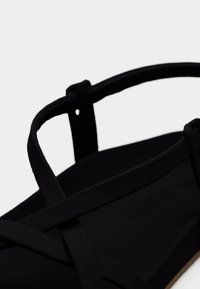 Μαύρα σανδάλια με μίνιμαλ σχεδιασμό, που διαθέτουν σταυρωτά λουριά, λείο ματ φινίρισμα και ανοιχτόχρωμη σόλα. Χωρίς ορατά μεταλλικά στοιχεία ή σχέδια.