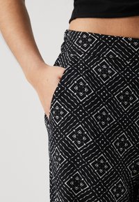 Person med svarta och vita mönstrade byxor, med handen i sidofickan, synlig midja och svart kort topp.