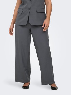 Femme portant un pantalon large gris foncé et un gilet sans manches boutonné assorti, associé à des ballerines noires pointues, debout sur fond blanc.