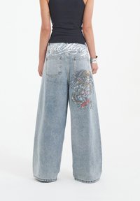 Jasnoniebieskie dżinsy o szerokich nogawkach mają haftowany wzór smoka w żywych kolorach na tylnej kieszeni. Wysoki stan z graficznym pasem.