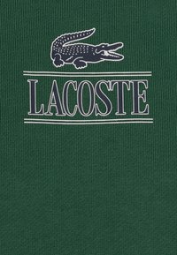Textil de rayas verde con un prominente logo de cocodrilo blanco y marino y el texto "LACOSTE" encima, creando una superficie texturizada.