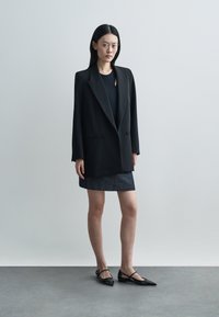 Sort dobbeltradet blazer med skarpe revers, båret over en sort top og en mørk denimnederdel, kombineret med spidse sorte flade sko.