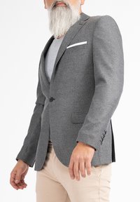 Blazer grigio con texture, dal taglio su misura, chiusura a un bottone, colletto a bavero e fazzoletto bianco, abbinato a pantaloni beige chiaro.