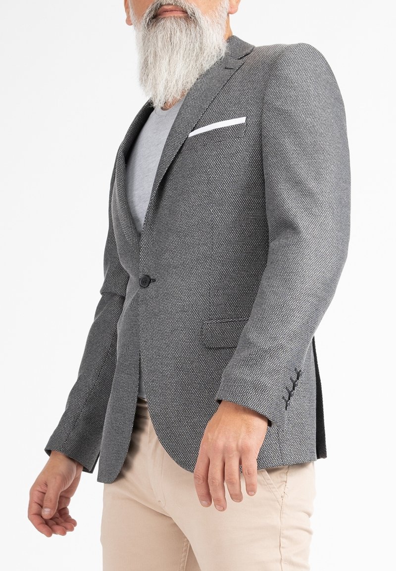 Blazer grigio con texture, dal taglio su misura, chiusura a un bottone, colletto a bavero e fazzoletto bianco, abbinato a pantaloni beige chiaro.