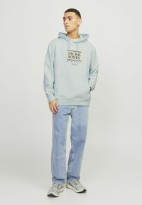Jasnoniebieska bluza z kapturem z kieszenią z przodu i graficznym logo, w zestawieniu z jasnoniebieskimi dżinsami i szarymi sneakersami. Miękka tkanina i luźny krój.