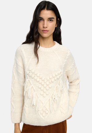 Kobieta z długimi ciemnymi włosami, ubrana w kremowy sweter z dzianiny o fakturowanych wzorach, z frędzlami oraz brązowe spodnie, na jednolitym tle.