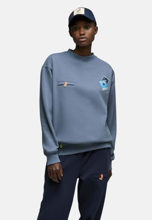 Personne portant un sweat-shirt bleu avec des patchs photo et un pantalon bleu marine avec des poches à languettes orange, assorti d'une casquette de baseball bleu marine avec un patch flamant rose.